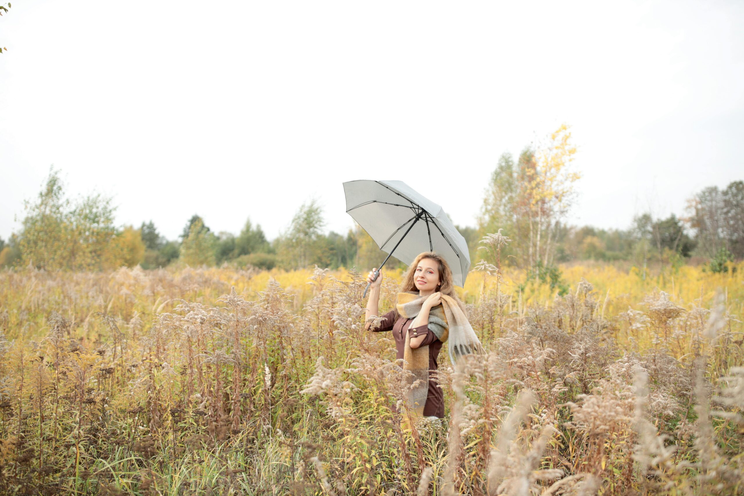 Eine Frau steht mit einem Regenschirm in einer herbstlichen Wiese, umgeben von Herbstlaub.